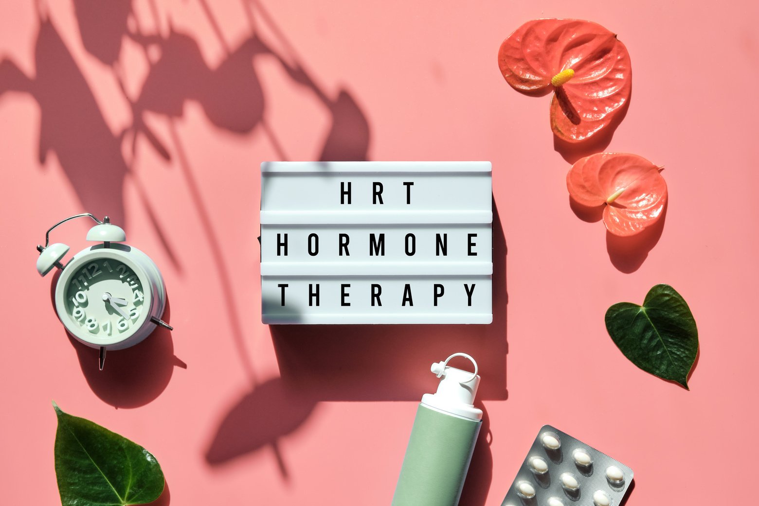 Text HRT Replacement  on Light Box. Menopause, Hormone  Concept. Pink Background with Alarm Clock, Exotic Leaves, Pills, Estrogene Gel.